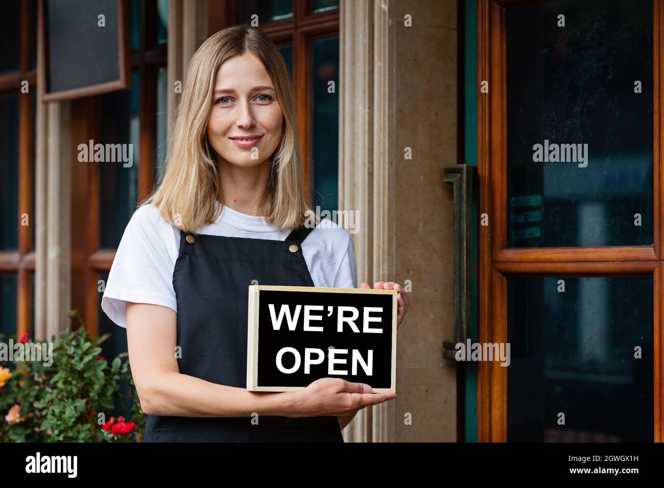 Jeune femme caucasienne de 30 ans avec des cheveux blond courts tenant une pancarte ouverte près des portes du café. Rouvrir le restaurant après le coronavirus Covid-19 Banque D'Images