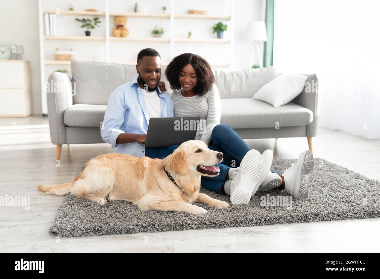 Concept de vie domestique. Portrait de famille noire aimante heureuse assise sur tapis de sol avec chien, en utilisant l'ordinateur et regarder le film, la navigation internée Banque D'Images