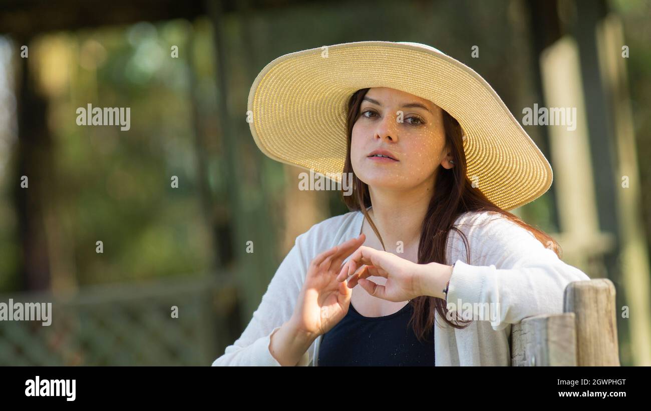 Portrait d'une belle jeune femme hispanique à cheveux longs portant un chapeau assis sur un banc de parc avec une attitude pensive contre un fond de non focalisé Banque D'Images