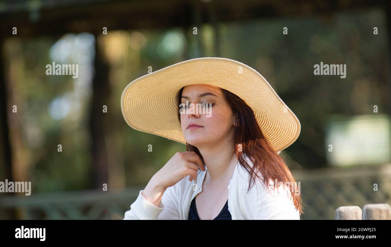 Portrait d'une belle jeune femme hispanique à cheveux longs portant un chapeau assis sur un banc de parc avec une attitude pensive contre un fond de non focalisé Banque D'Images