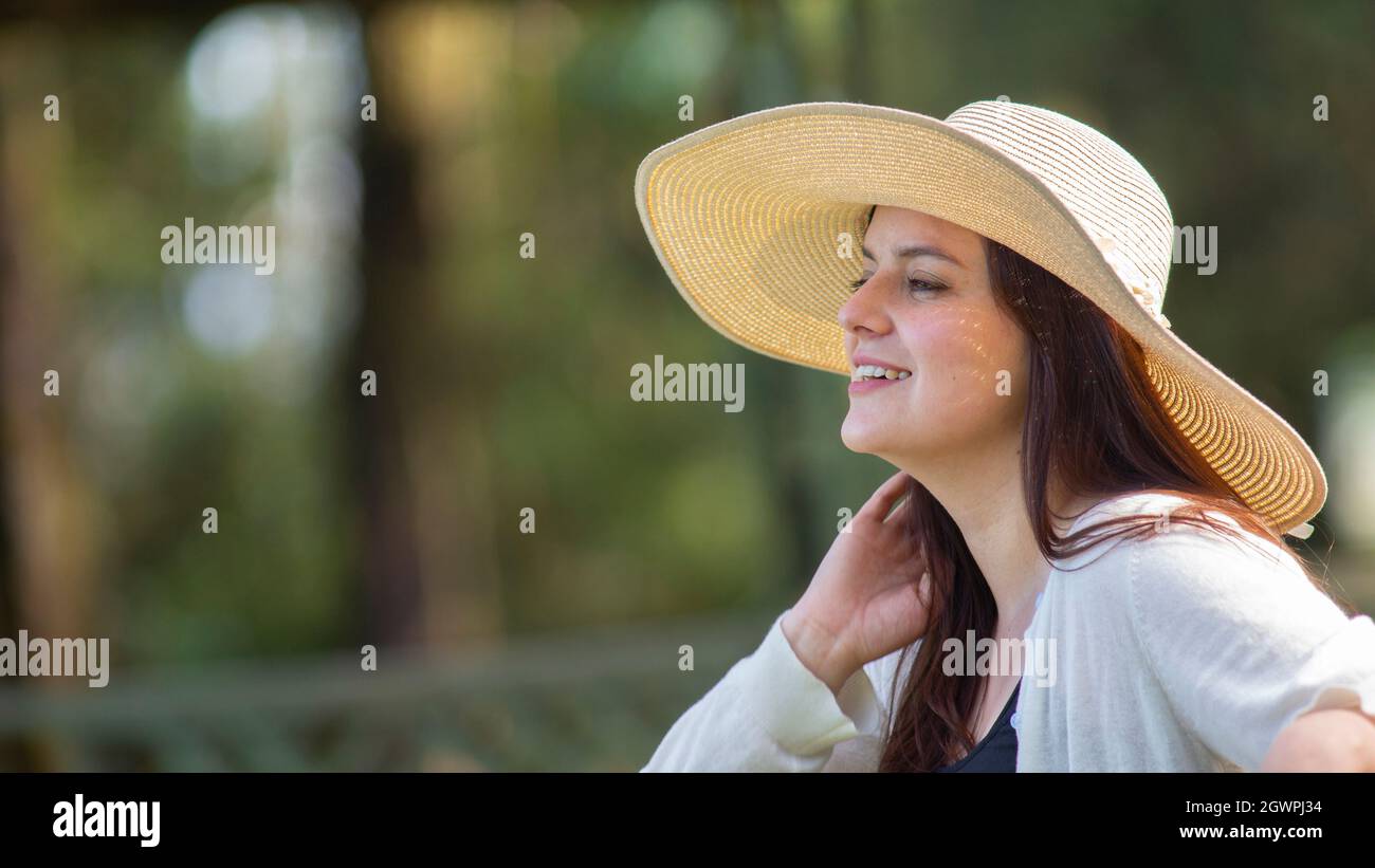 Portrait d'une belle jeune femme hispanique aux cheveux longs avec un chapeau très gai assis sur un banc de parc sur un fond de votre vert non concentré Banque D'Images