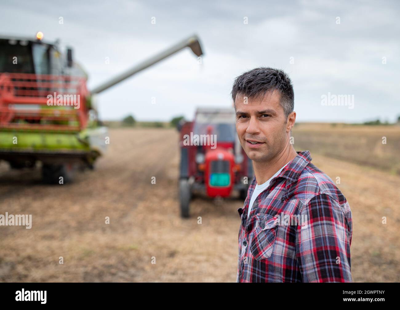 Jeune agronome attrayant debout dans le champ de récolte regardant la caméra. Moissonneuse-batteuse remplissant la remorque du tracteur de grains Banque D'Images
