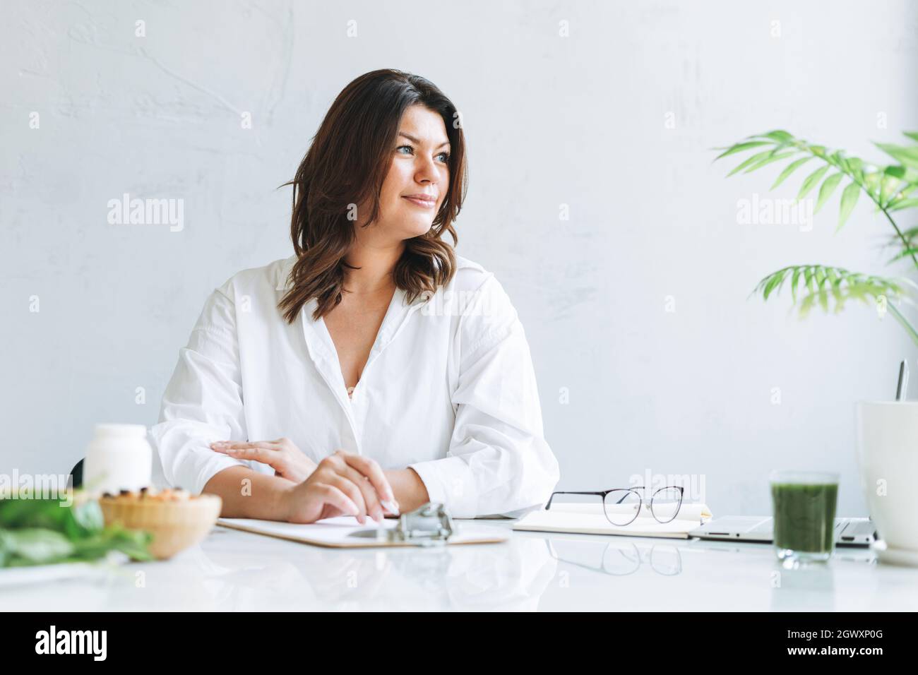 Portrait de jeune femme brunette souriante médecin nutritionniste plus taille en chemise blanche travaillant à l'ordinateur portable dans une salle de bureau moderne et lumineuse. Le médecin préc Banque D'Images