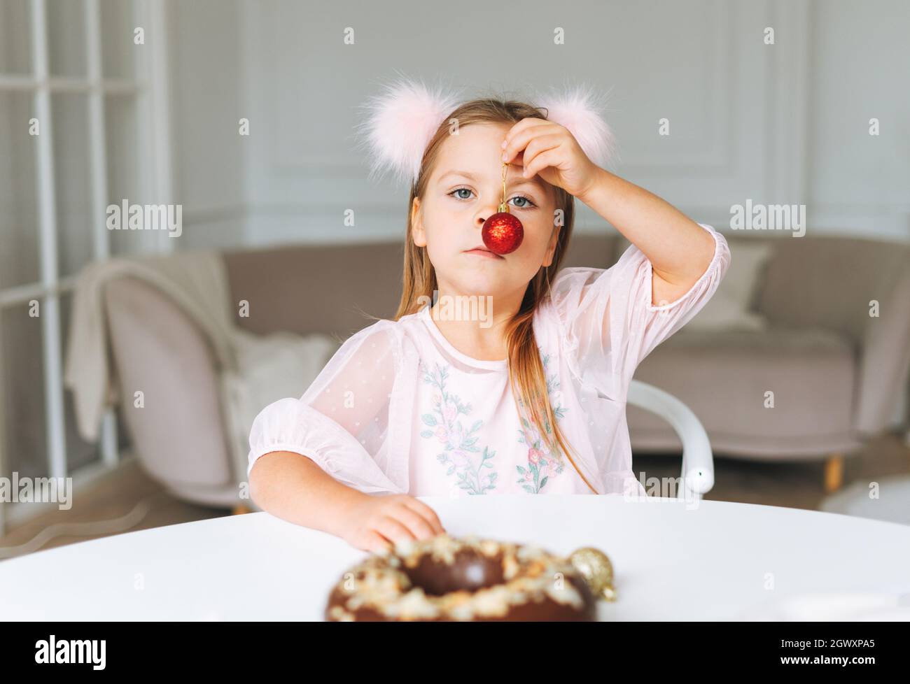Drôle petite fille mignonne avec de longs cheveux dans la robe rose clair avec boule de noël rouge dans les mains dans le salon lumineux à la maison. Noël, anniversaire Banque D'Images