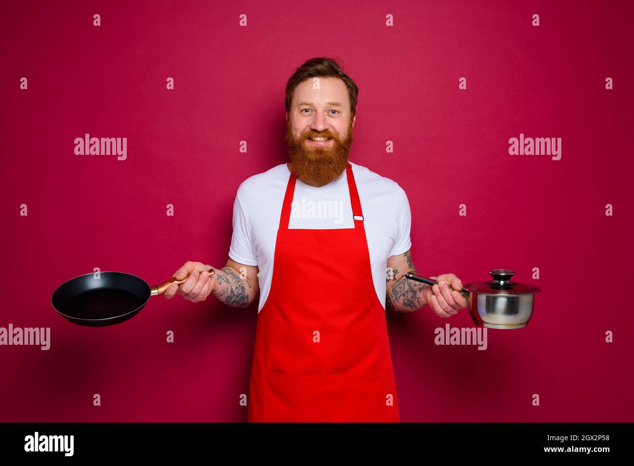 Un chef heureux avec une barbe et un tablier rouge cuisiniers avec une casserole et une casserole Banque D'Images