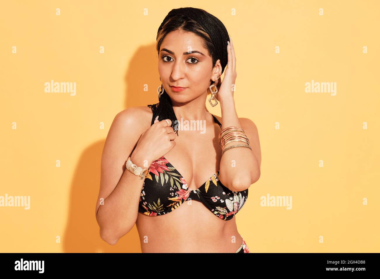 Peau naturelle sexy fille latine portant un bikini avec des fleurs. Elle porte des compléments de joailerie et elle pose devant un fond jaune regardant Banque D'Images