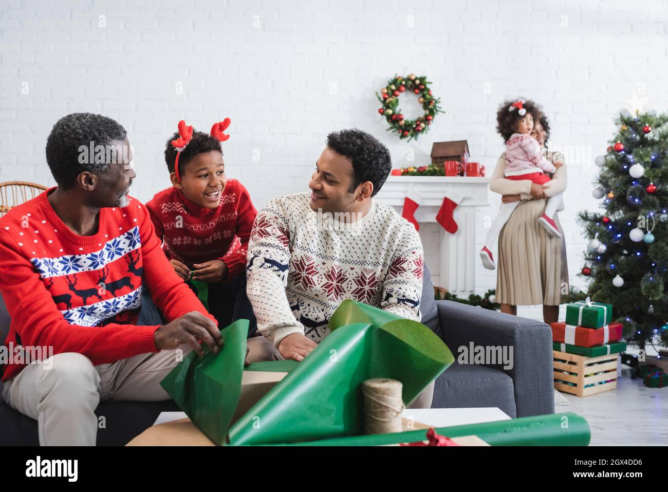 afro-américain garçon dans les cornes de renne serre-tête souriant près des hommes emballage cadeaux et flou mère avec enfant près de l'arbre de noël Banque D'Images