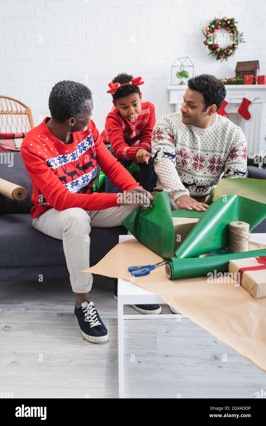 un garçon américain africain en corne de renne serre-tête pointant sur une table avec des boîtes-cadeaux et du papier d'emballage près de papa et grand-père Banque D'Images