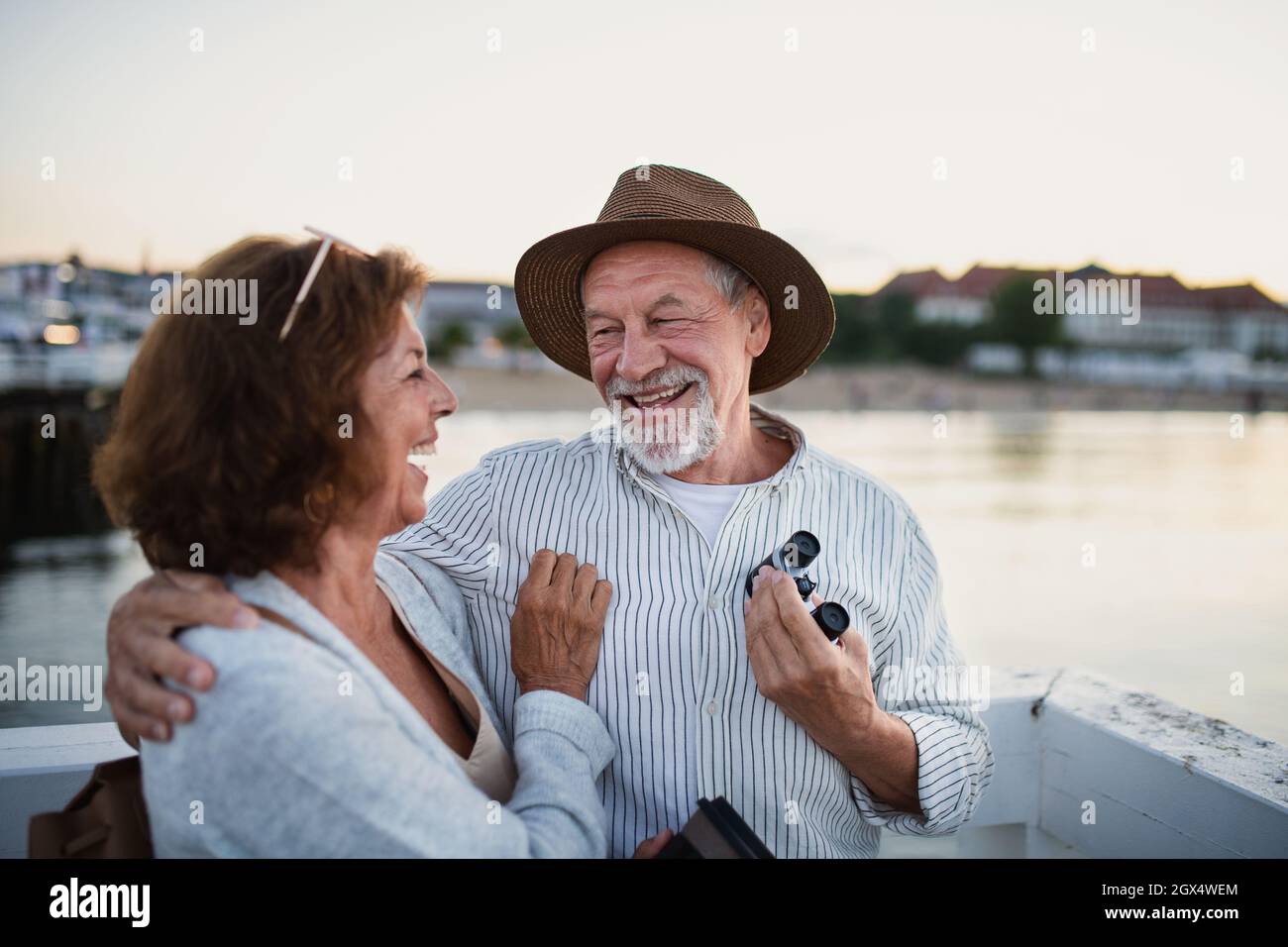 Un couple aîné heureux qui s'enserre à l'extérieur sur la jetée en bord de mer, en regardant chaque autre et en riant. Banque D'Images