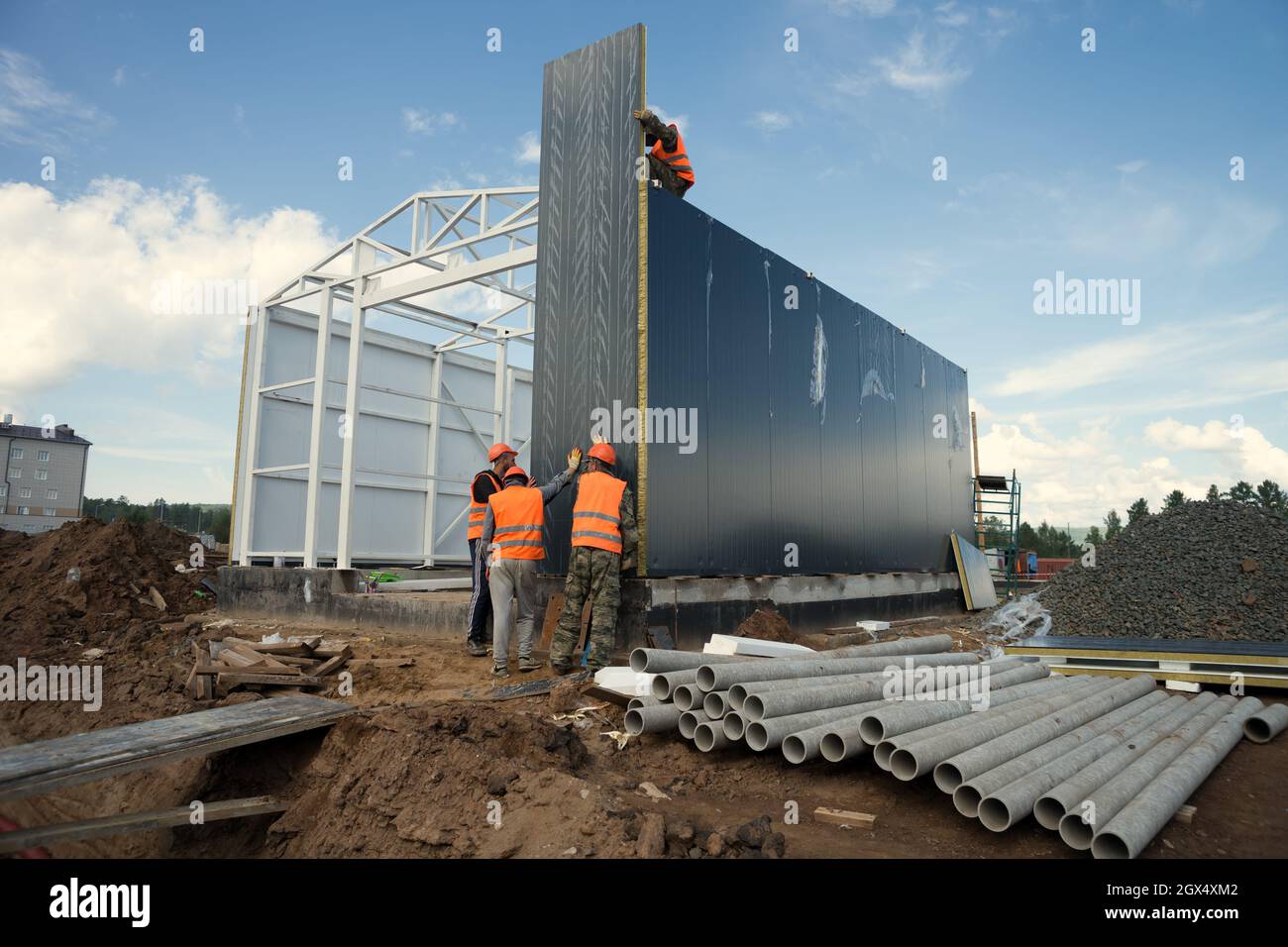 Les travailleurs en casque de sécurité installent un panneau mural sur le cadre d'un bâtiment industriel sur un chantier de construction en été. Banque D'Images