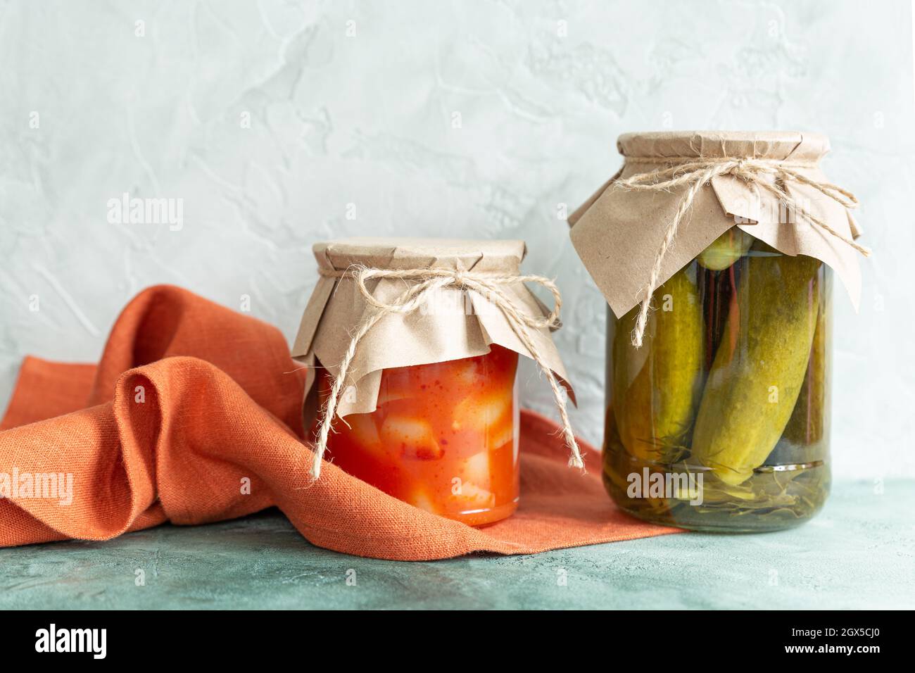 Cornichons, légumes en conserve en pots de verre et serviette en toile de terre cuite sur fond vert clair. Repas faits maison, stockage de nourriture. Produits écologiques emballés Banque D'Images