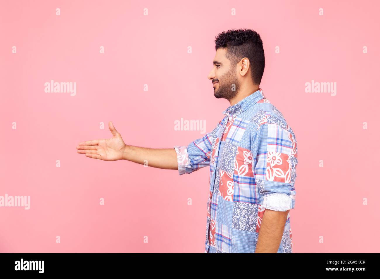 Vue latérale d'un sympathique homme barbu dans une chemise bleue de style décontracté, en se faisant main pour se serrer la main, en se familiarisant avec l'entrevue d'emploi, en rencontrant de nouvelles personnes. Studio d'intérieur isolé sur fond rose. Banque D'Images