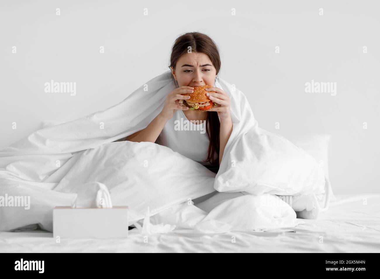 Une jeune femme européenne affamée s'assoit au lit à la maison et mange du hamburger Banque D'Images