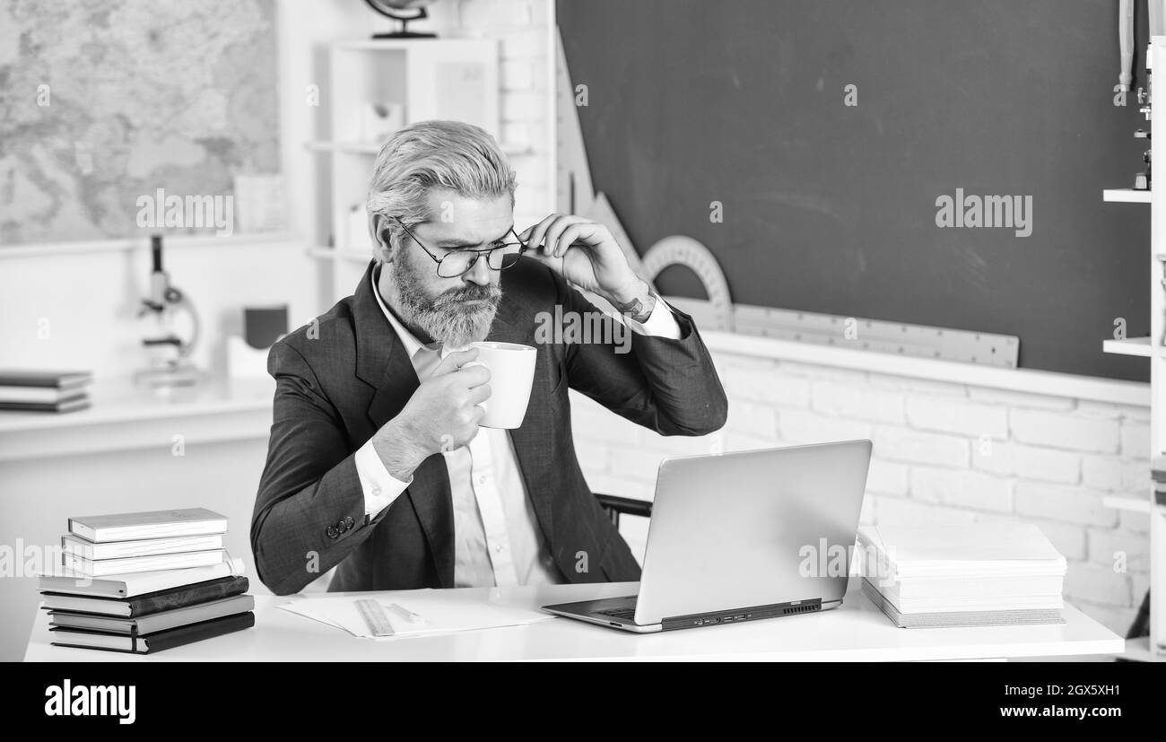 Cours en ligne. Professeur principal utilise l'ordinateur. Un professeur d'informatique boit du thé au bureau de l'école. Homme barbu travaille dans une salle de classe informatique. Nouvelle technologie Banque D'Images