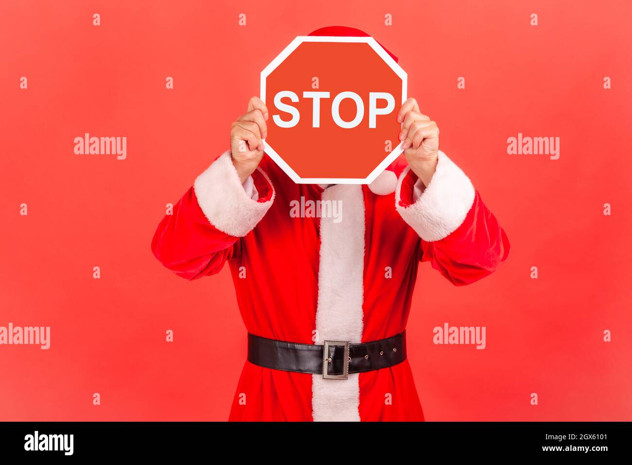 Homme inconnu portant le costume du père noël couvrant le visage avec le symbole Stop, personne anonyme tenant le panneau rouge de signalisation, avertissement à partir, concept d'interdiction. Studio d'intérieur isolé sur fond rouge. Banque D'Images