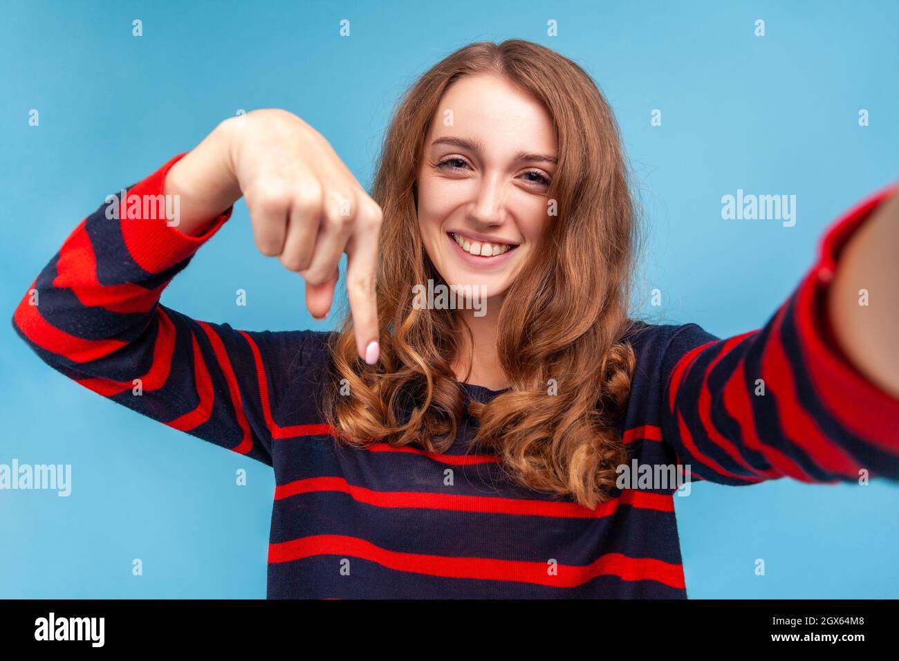 Femme souriante portant un pull rayé style décontracté, prenant selfie, point de vue de la photo, pointant le doigt vers le bas, recommander de souscrire son vlog. Studio d'intérieur isolé sur fond bleu. Banque D'Images