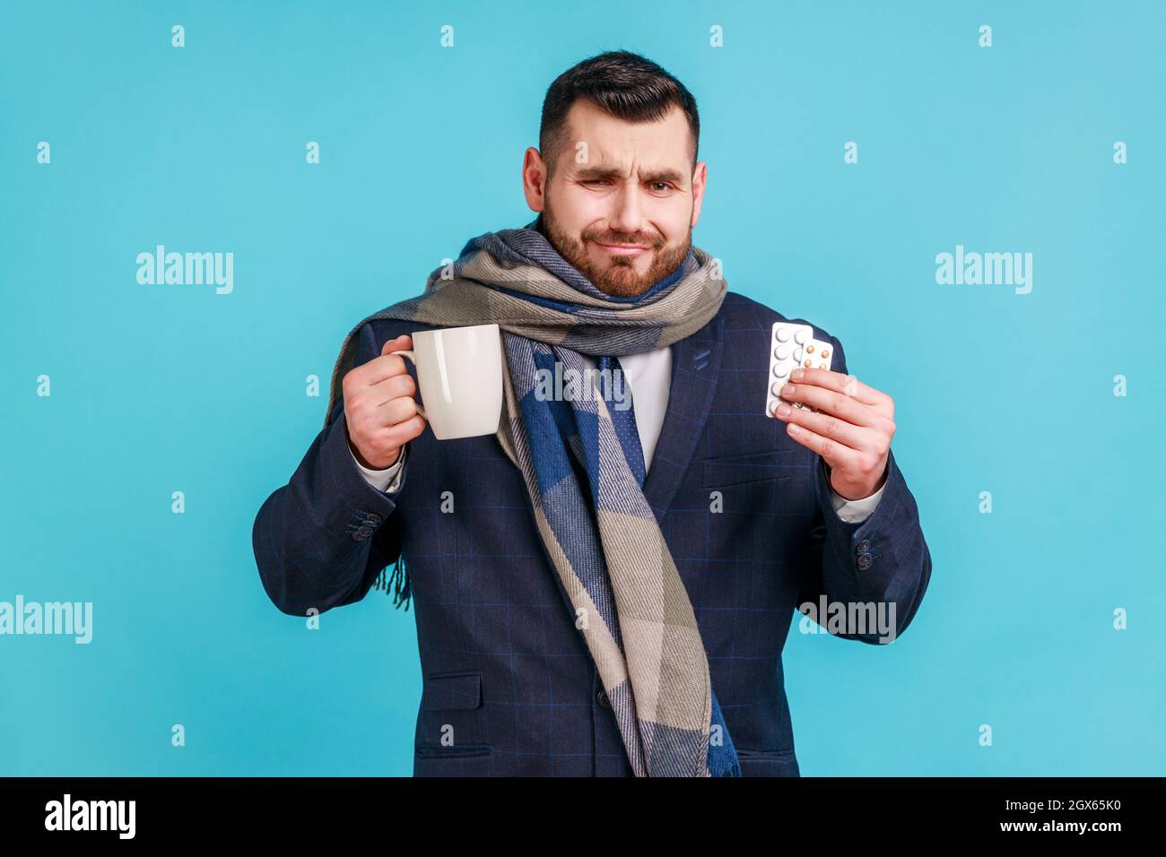 Portrait d'une triste entreprise malade portant un costume de style officiel et enveloppé dans un foulard, traitant avec des pilules et du thé chaud, étant bouleversé, se sentant mal. Studio d'intérieur isolé sur fond bleu. Banque D'Images