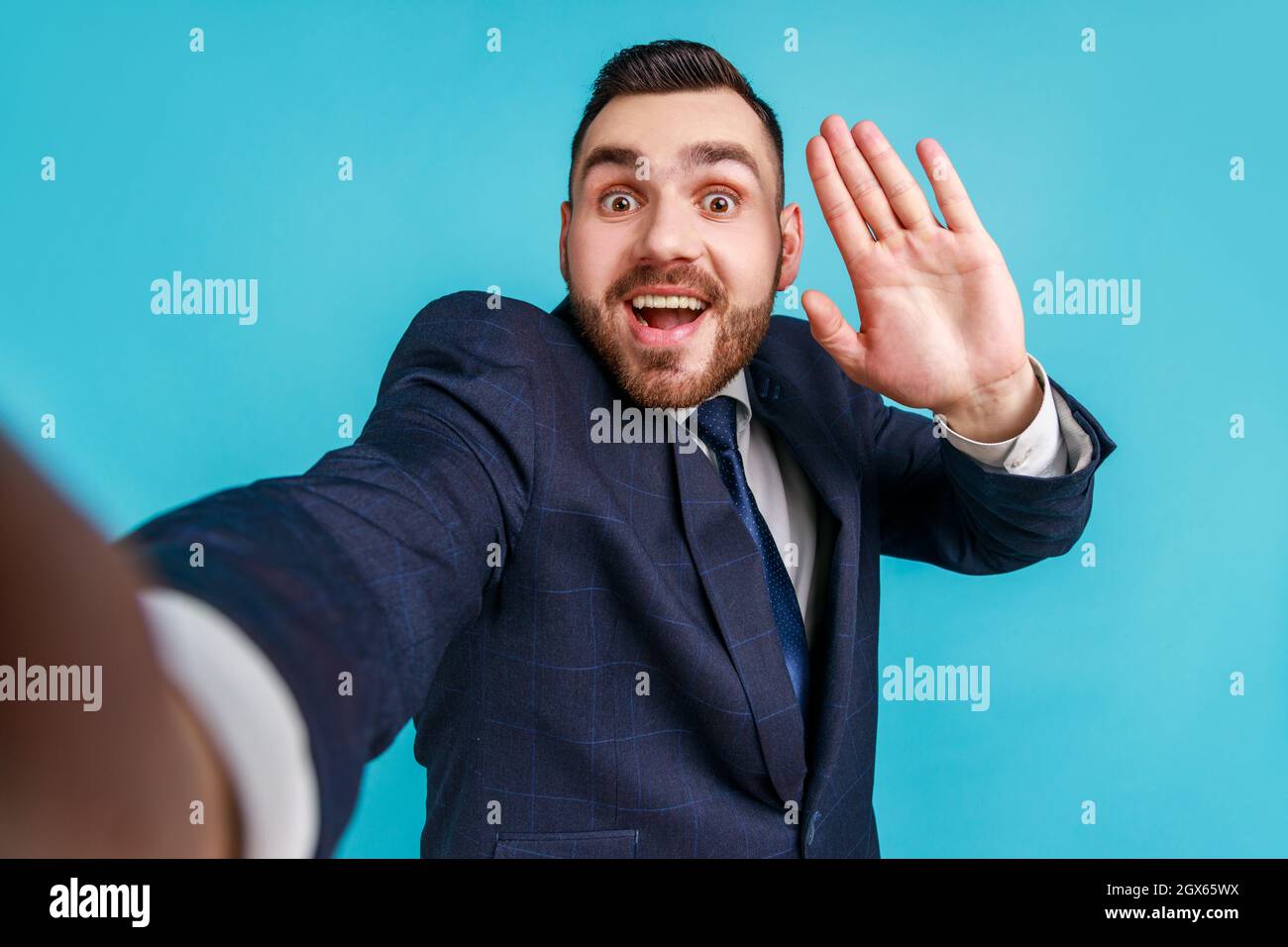 Jeune adulte enthousiaste satisfait homme portant un costume de style officiel, faisant selfie ou streaming, en agitant main POV, point de vue de la photo. Studio d'intérieur isolé sur fond bleu. Banque D'Images