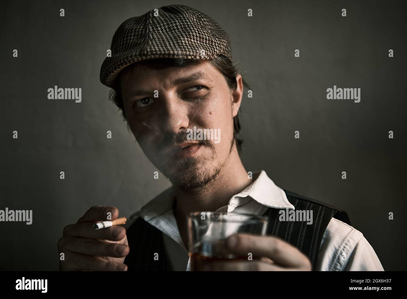 Un jeune homme fume une cigarette et boit du whisky, souffle de la fumée, vêtu d'un style rétro, dans un béret, une prise de vue cinématographique, un gros plan Banque D'Images