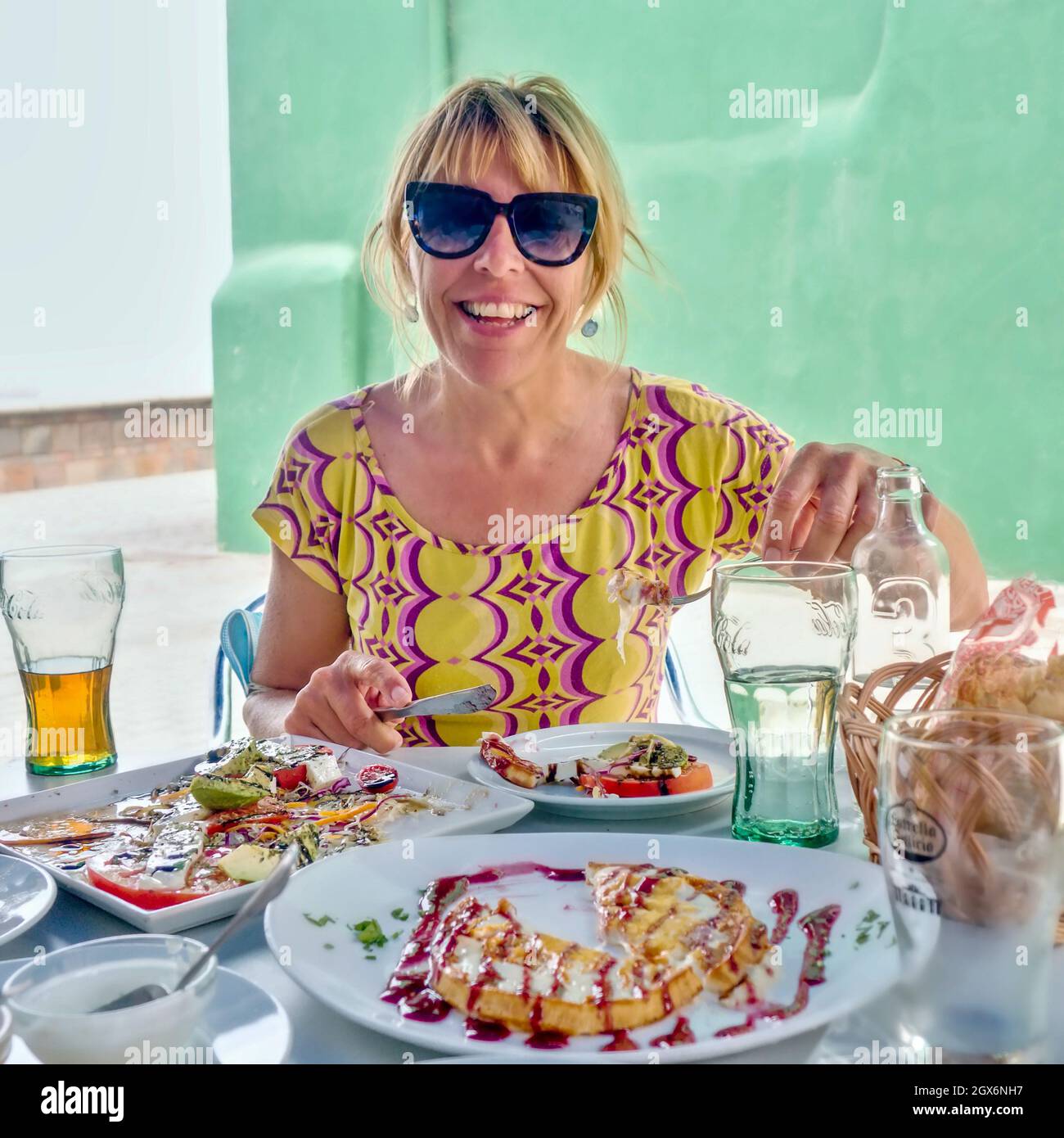 une jeune femme avec des lunettes de soleil et des cheveux blonds brochés est assise à une table richement fixée avec un repas avec de la nourriture et des boissons à l'extérieur sur une terrasse. Banque D'Images