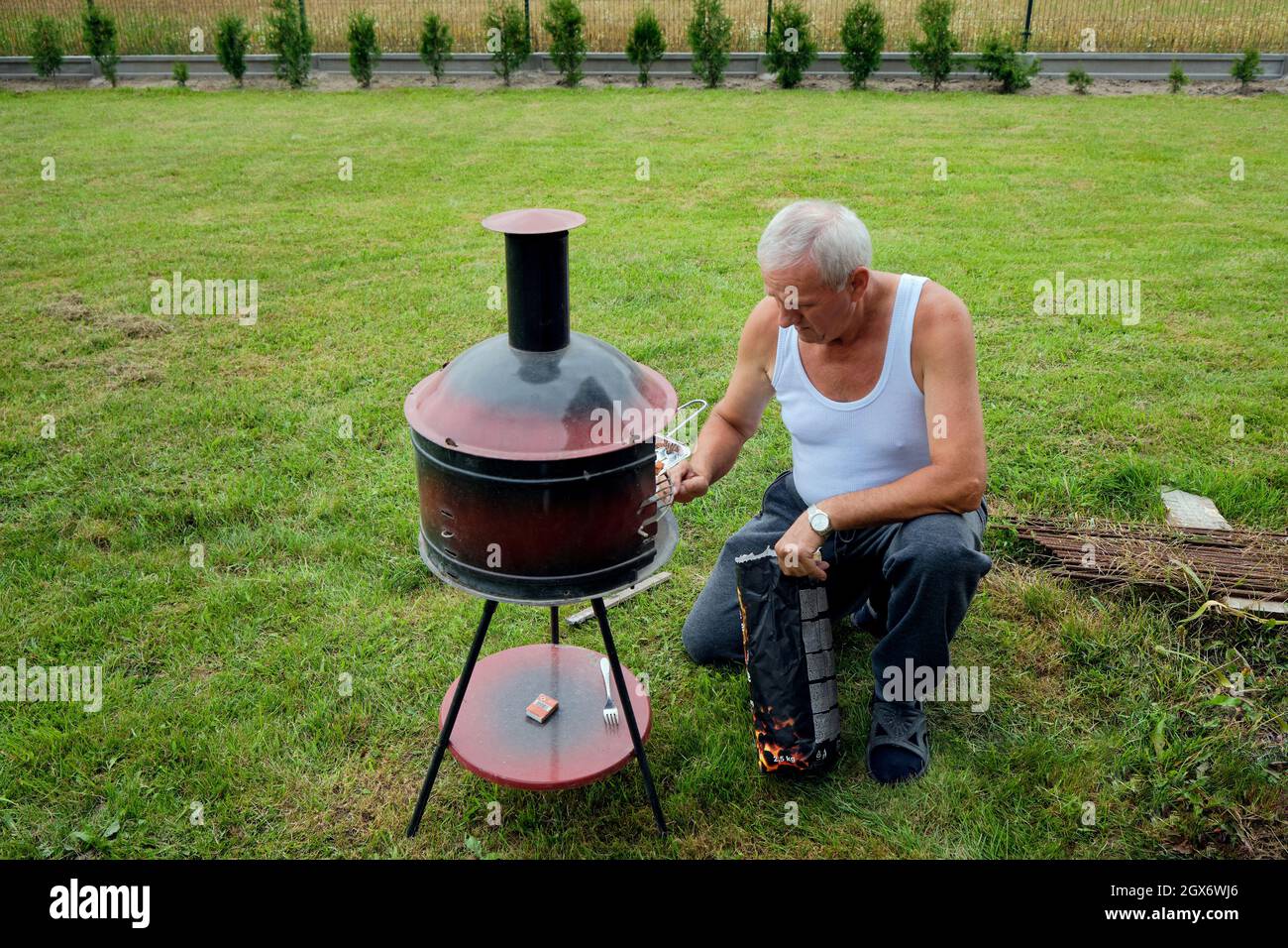 un homme âgé s'agenouille à un barbecue à l'extérieur sur fond d'herbe verte Banque D'Images