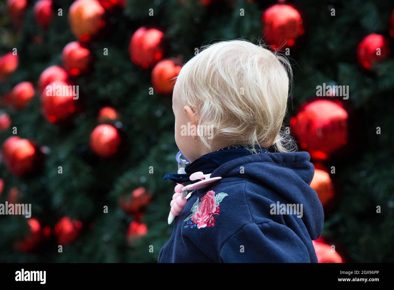 Blonde blanche bébé fille en bleu foncé pull à capuche avec fleur rouge regarder l'arbre de Noël avec de grosses balles de nouvel an.Vacances d'hiver pour les enfants Banque D'Images