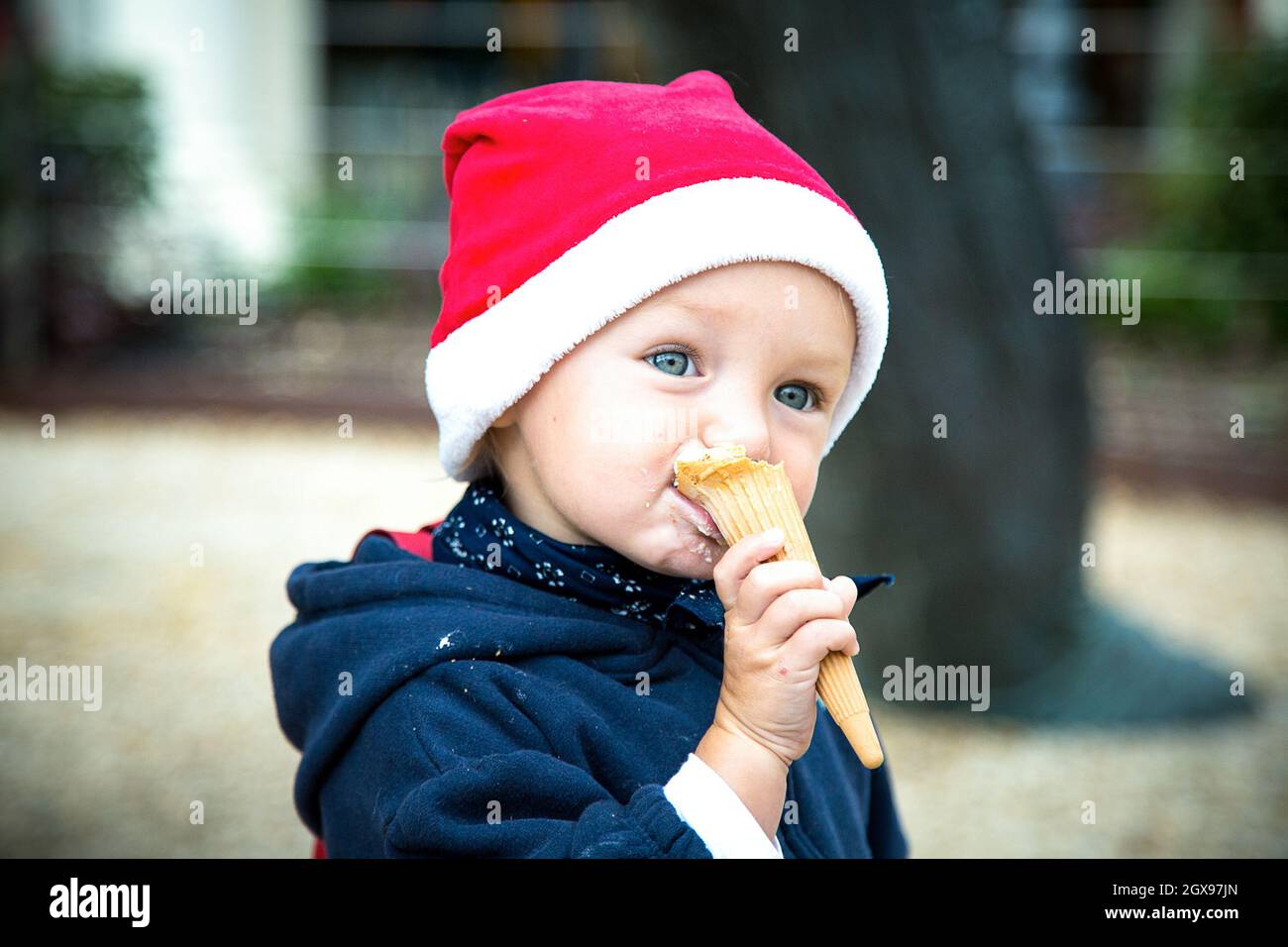 Bleu oeil bébé fille en manteau d'hiver bleu foncé et chapeau de Noël rouge mange de la glace dans le cône de gaufré.Un enfant de race blanche en vacances du nouvel an.Manger de la glace Banque D'Images