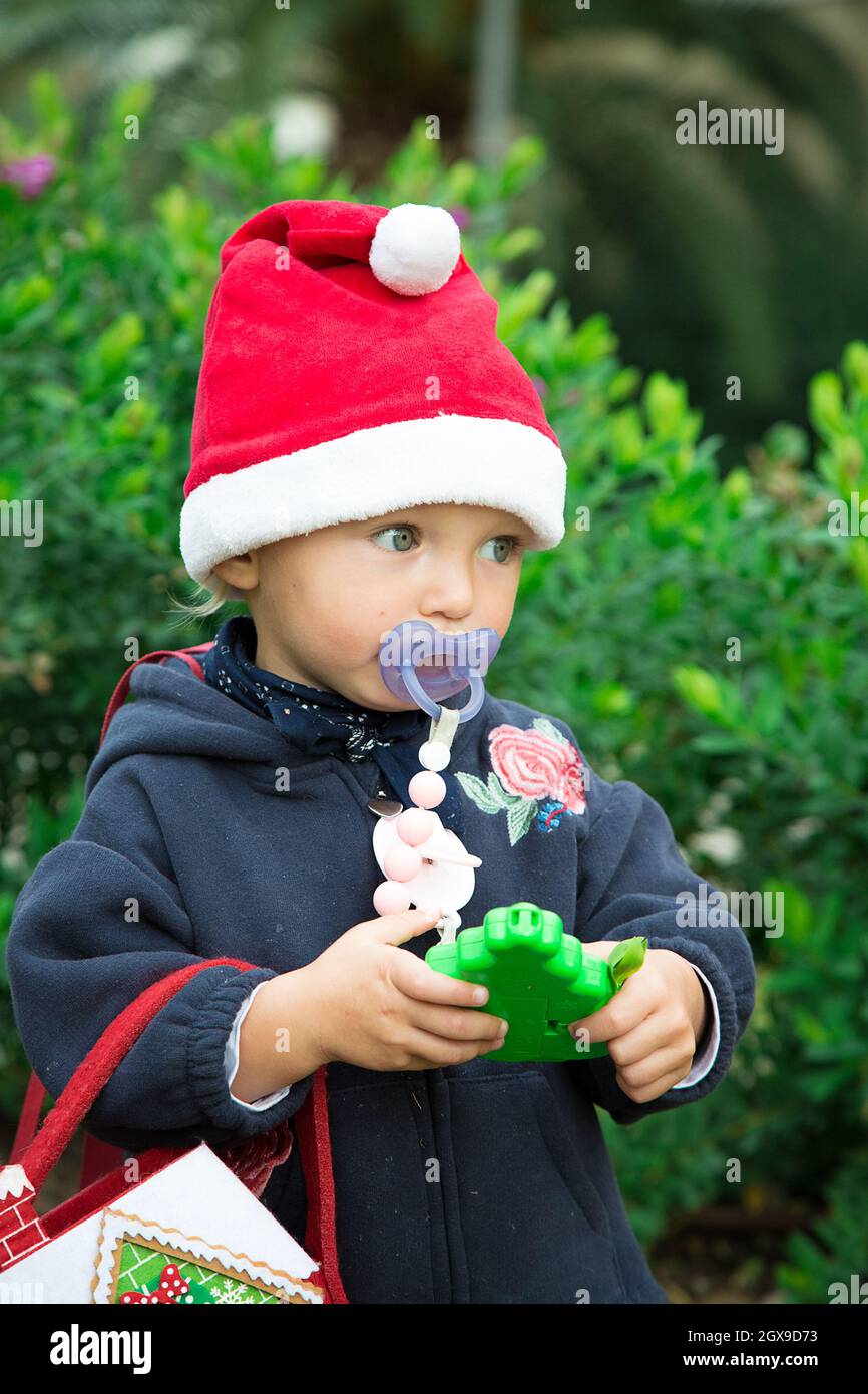Petite fille habillée pour Noël marche dehors avec des arbres verts derrière.Chapeau rouge, manteau bleu, sac en feutre, jouet vert dans les mains.Arbre du nouvel an.Vacances Banque D'Images