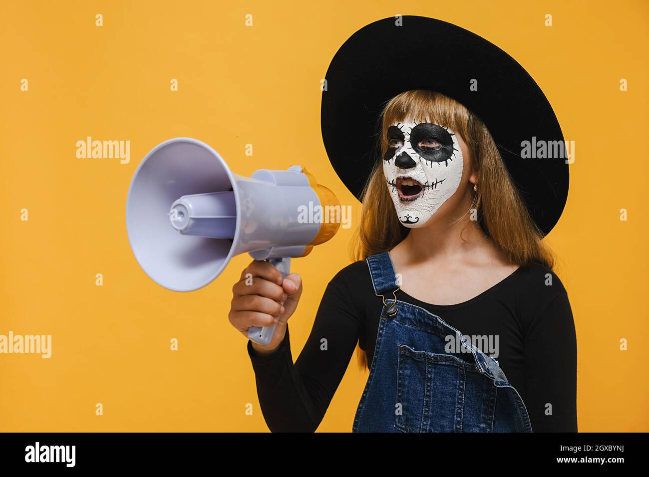 Portrait de l'enfant fille effrayante avec Halloween masque de maquillage hurlant dans le mégaphone regardant de côté, porte grand chapeau noir, posant isolé sur le mur jaune Banque D'Images
