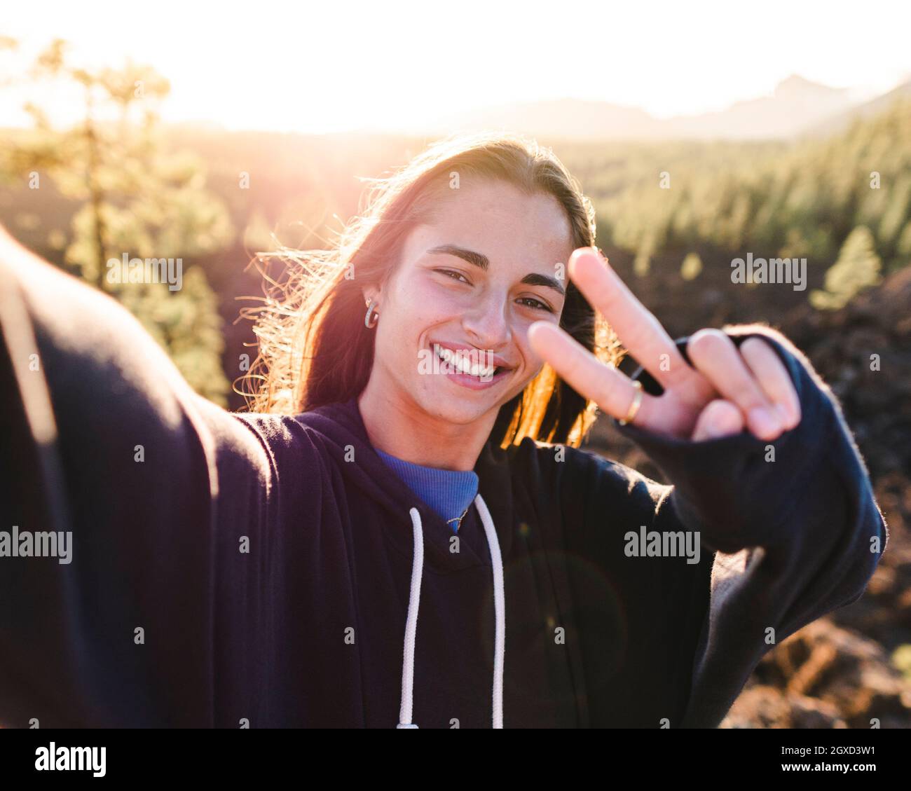 Une adolescente en sweat-shirt prend un selfie avec un smartphone tout en démontrant un geste de victoire en regardant l'appareil photo contre le support de Tenerife à l'arrière Banque D'Images