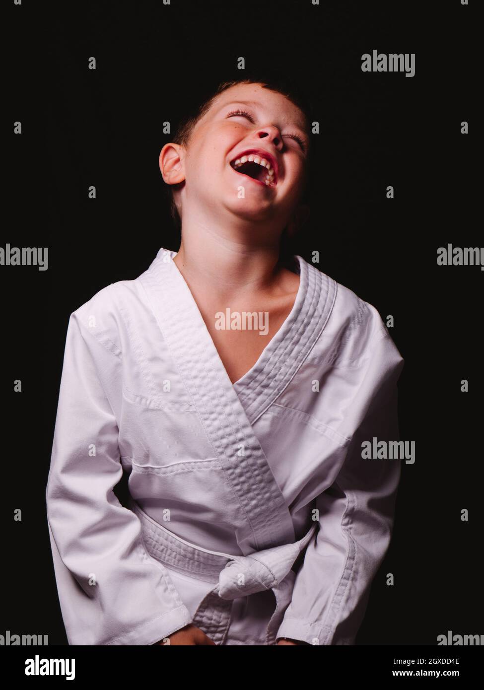 Mignon garçon dans un kimono karaté riant avec joie avec la bouche ouverte en studio sur fond noir Banque D'Images