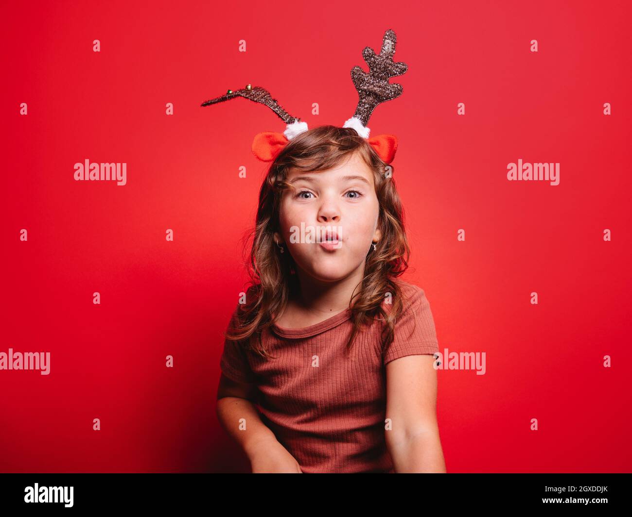 Joyeuse petite fille dans des vêtements décontractés et serre-tête cerf de fête soufflant baiser regardant l'appareil photo pendant la fête de Noël sur fond rouge Banque D'Images