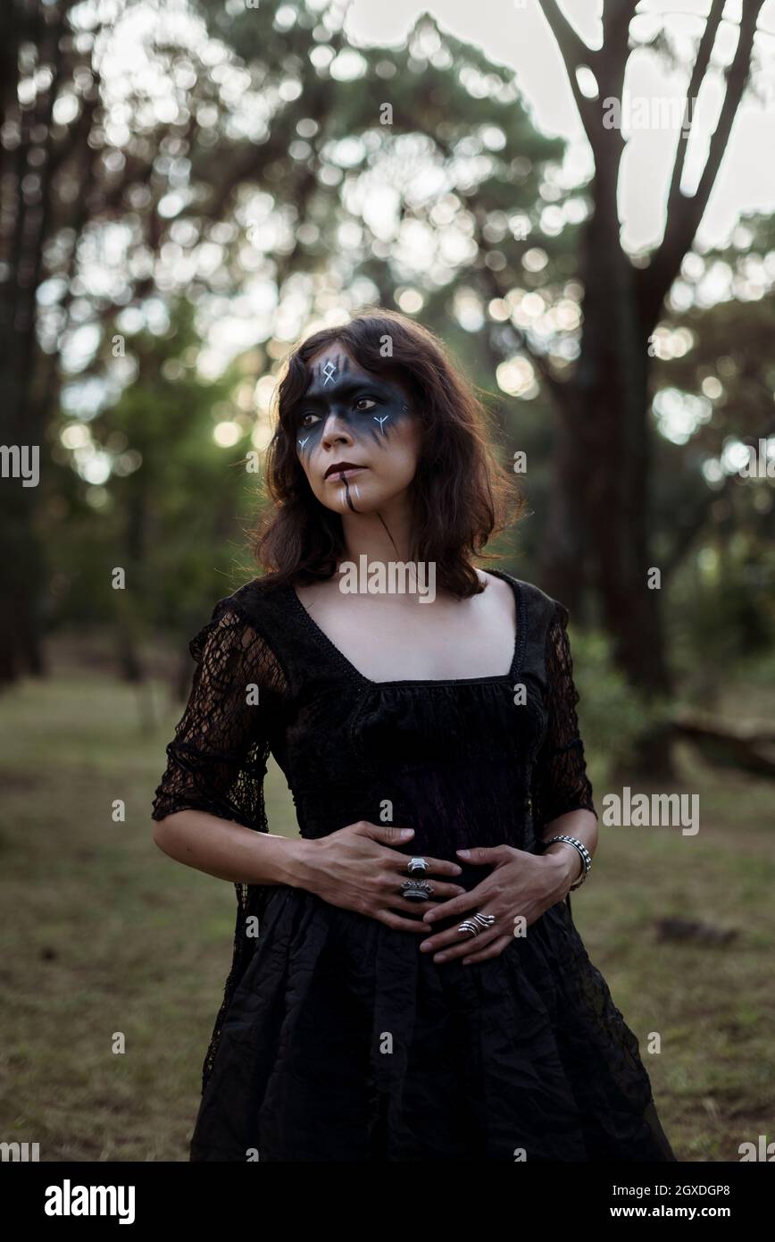 Sorcière mystique dans une longue robe noire et avec le visage peint se tenant à l'écart dans les bois sombres lugubres Banque D'Images