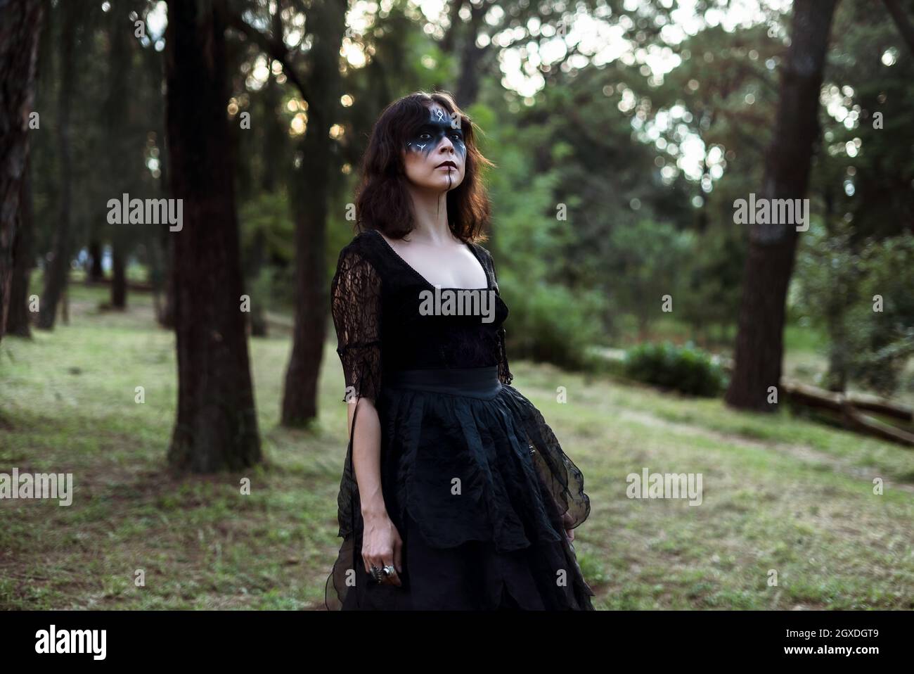 Sorcière mystique dans une longue robe noire et avec le visage peint se tenant à l'écart dans les bois sombres lugubres Banque D'Images