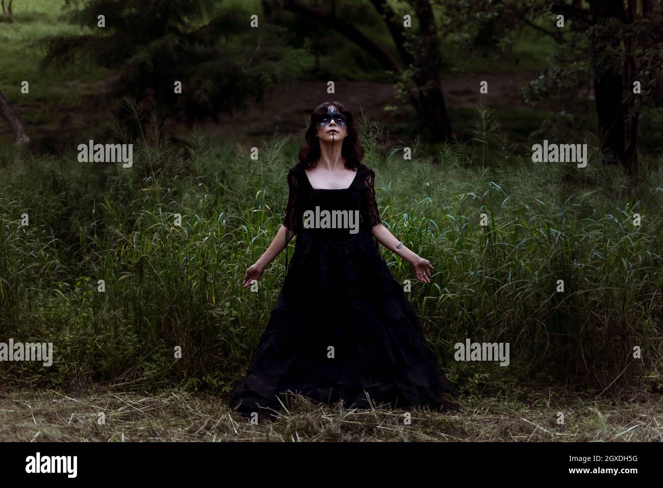 Sorcière mystique dans une longue robe noire et avec le visage peint debout regardant dans les bois sombres lugubres Banque D'Images