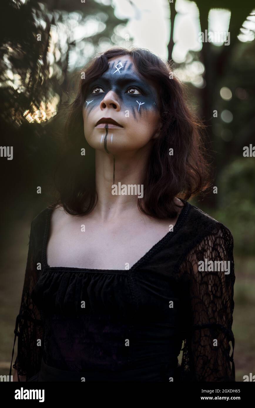 Sorcière mystique dans une longue robe noire et avec le visage peint debout regardant dans les bois sombres lugubres Banque D'Images