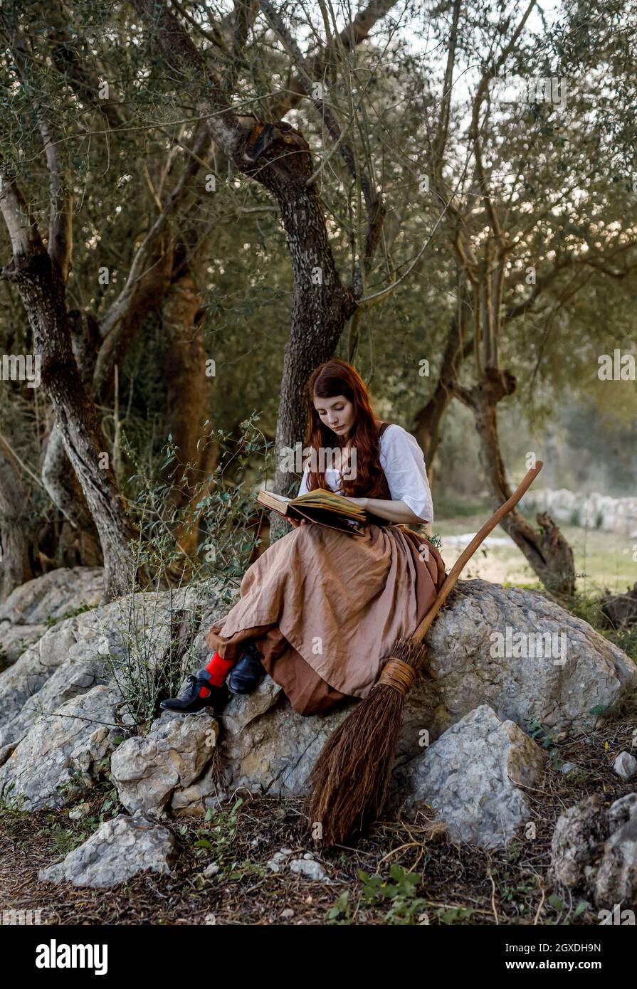 Une femme ciblée lisant un livre magique tout en étant assise sur un rocher près d'un balai dans les bois Banque D'Images