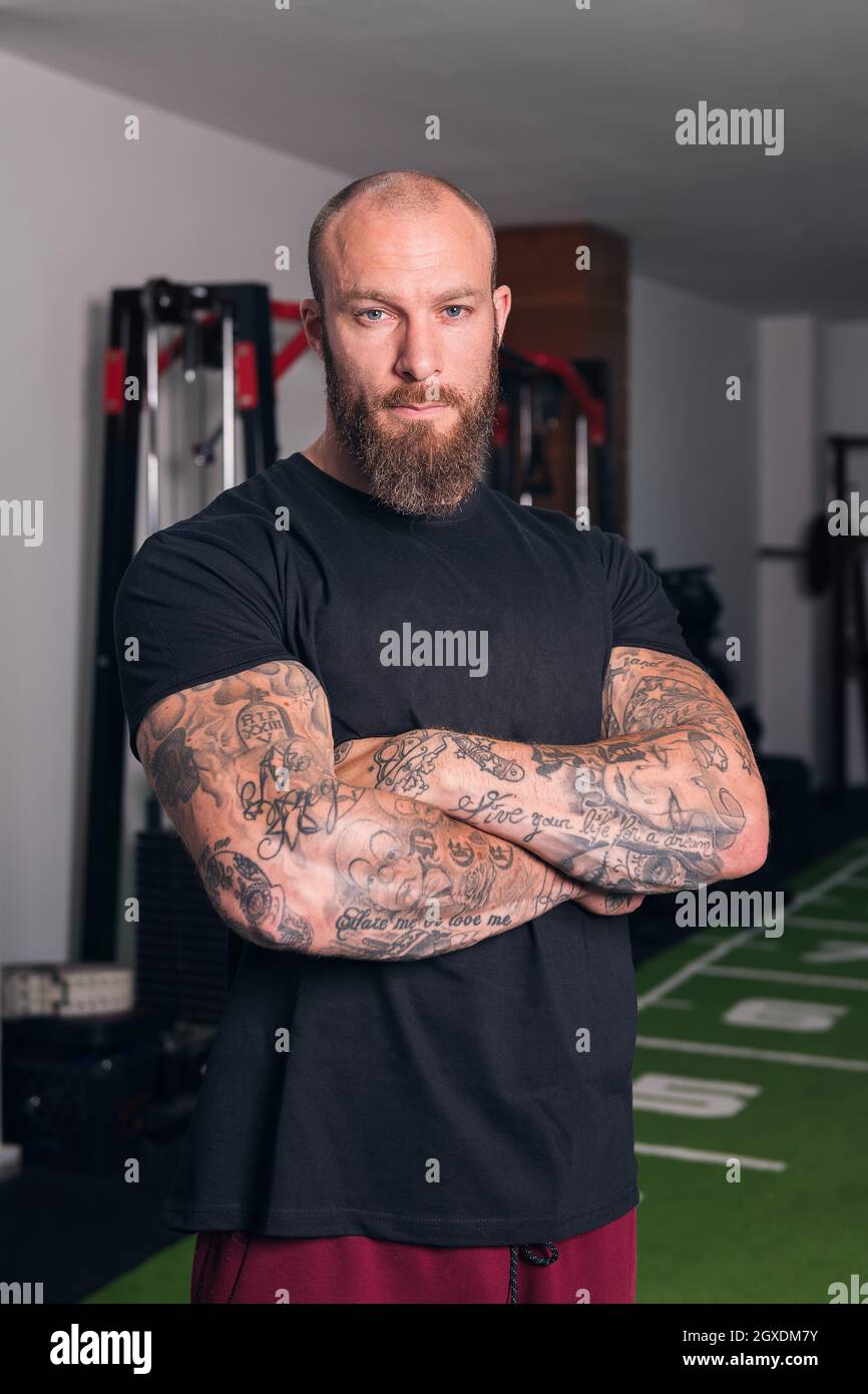 Sportif adulte fort avec barbe et tatouages sur les bras pliés regardant la caméra dans le gymnase Banque D'Images