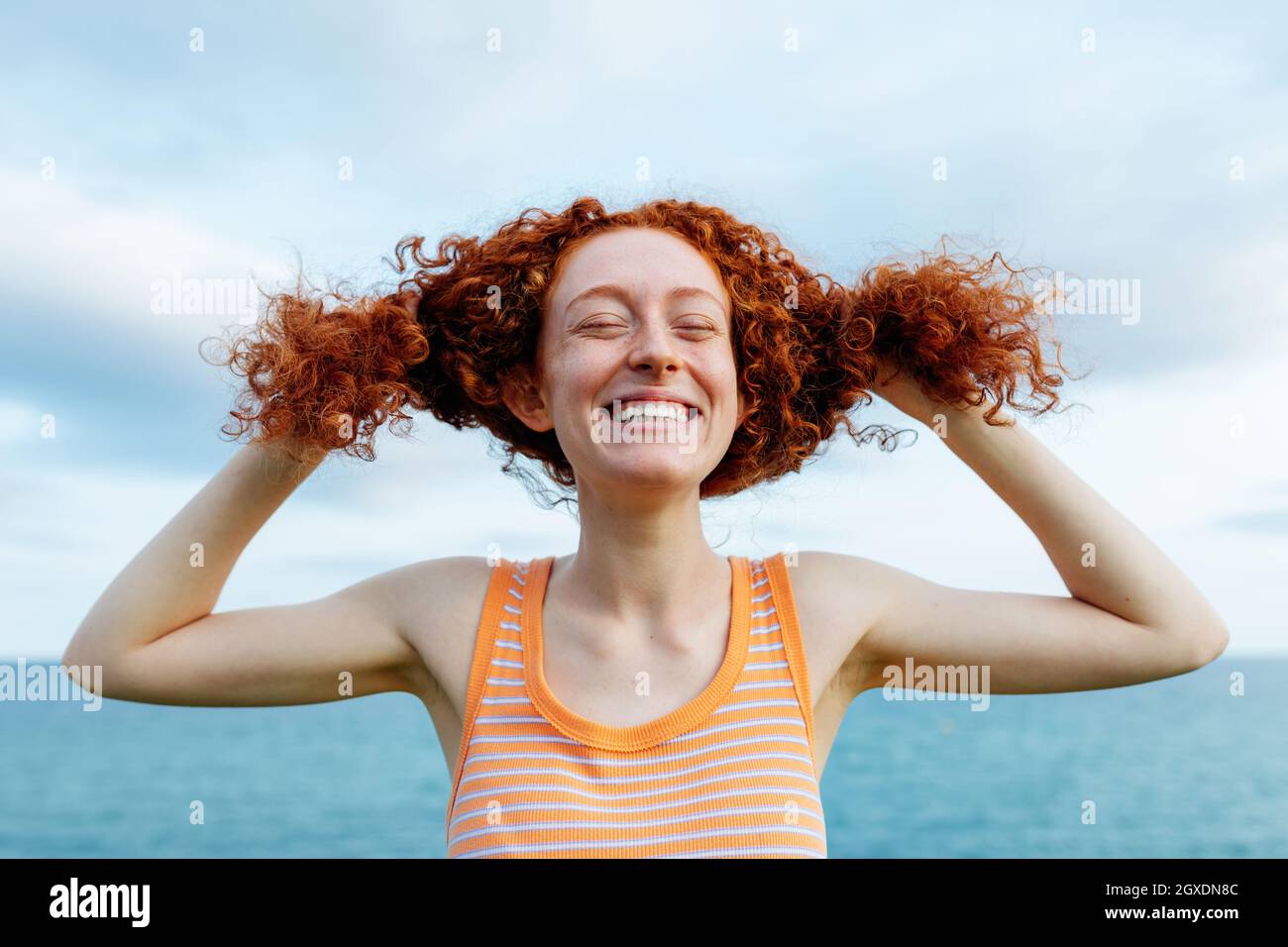 Joyeuse jeune femme à tête rouge faisant des queues de cheval avec des cheveux bouclés tout en profitant de la liberté sur la mer Banque D'Images