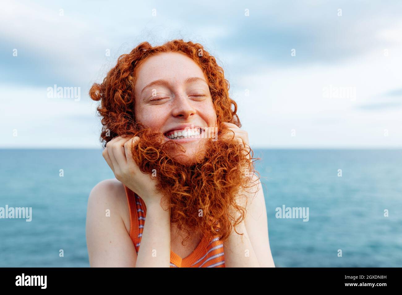 Joyeuse jeune femme à tête rousse faisant une pose puéril avec des cheveux bouclés tout en profitant de la liberté sur la mer avec les yeux fermés Banque D'Images