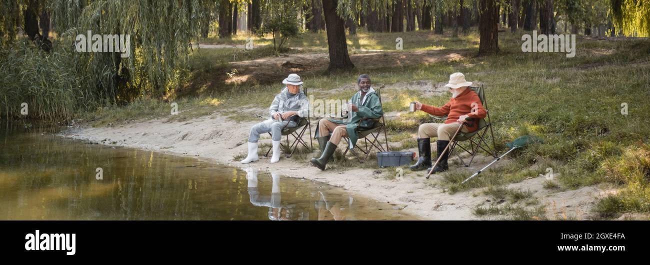 Homme afro-américain souriant tenant une tasse près d'amis interraciaux près du lac dans le parc, bannière Banque D'Images