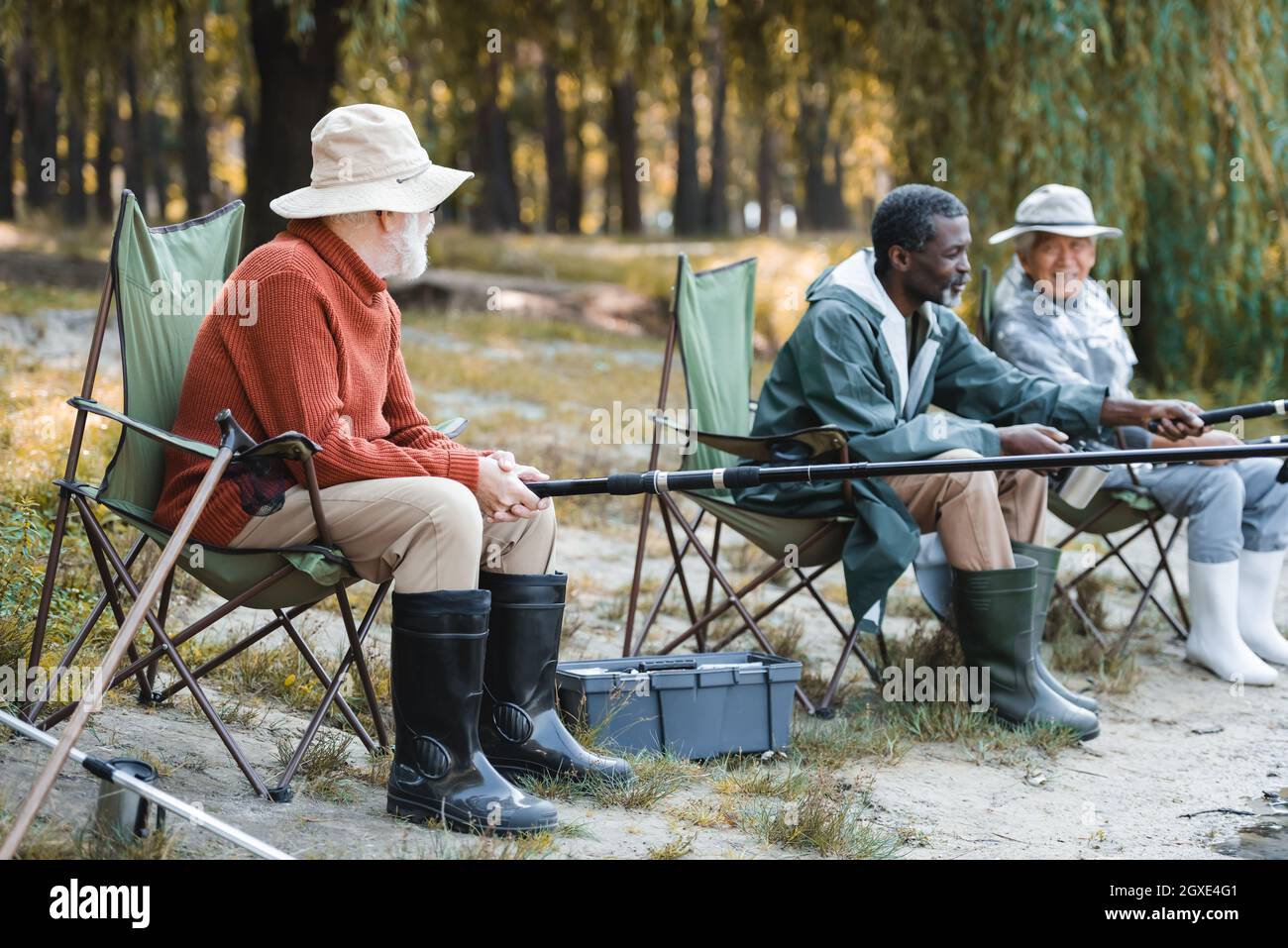 Homme âgé avec canne à pêche assis près de la boîte à outils et amis interraciaux à l'extérieur Banque D'Images