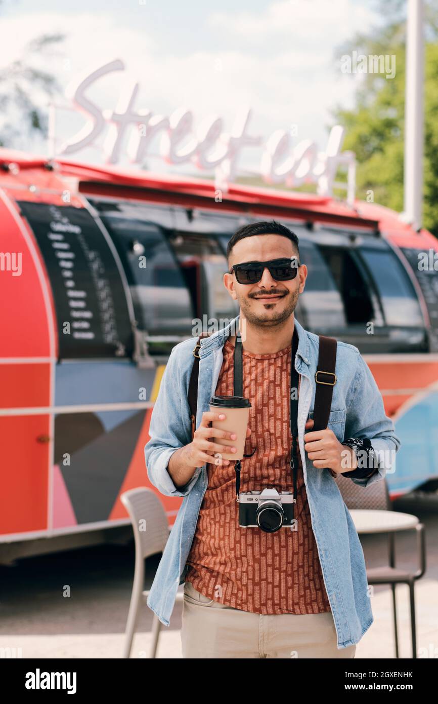 Un jeune photographe heureux avec une boisson et une photocamera debout contre un camion de nourriture de rue Banque D'Images