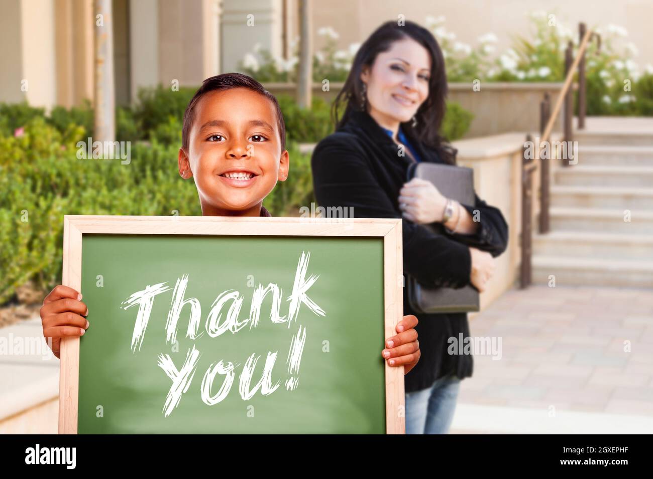 Happy Hispanic Boy Holding Thank You Chalk Board à l'extérieur sur le campus de l'école comme l'enseignant regarde sur. Banque D'Images