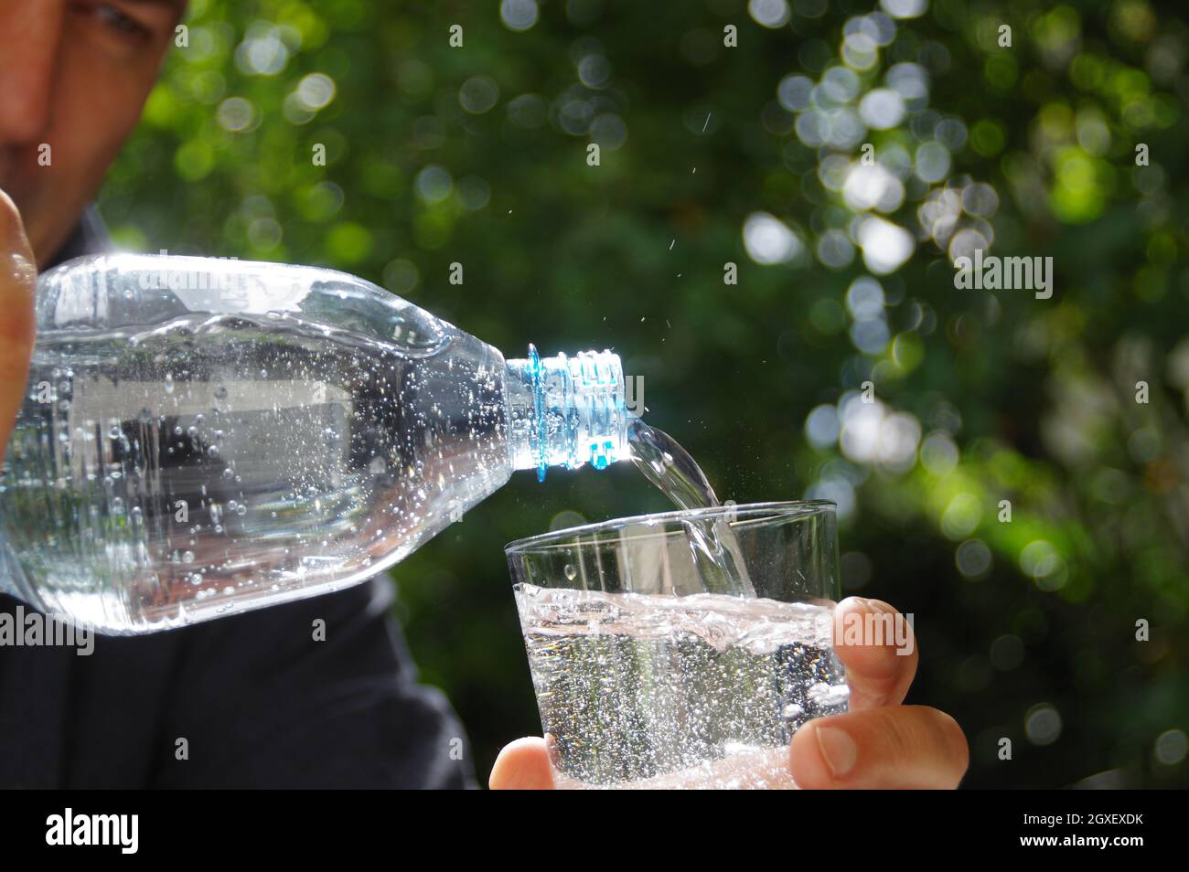 Eau versée dans un verre à la main avec le jardin en arrière-plan. Rafraîchissement avec de l'eau minérale propre naturelle dans une bouteille en plastique. Banque D'Images