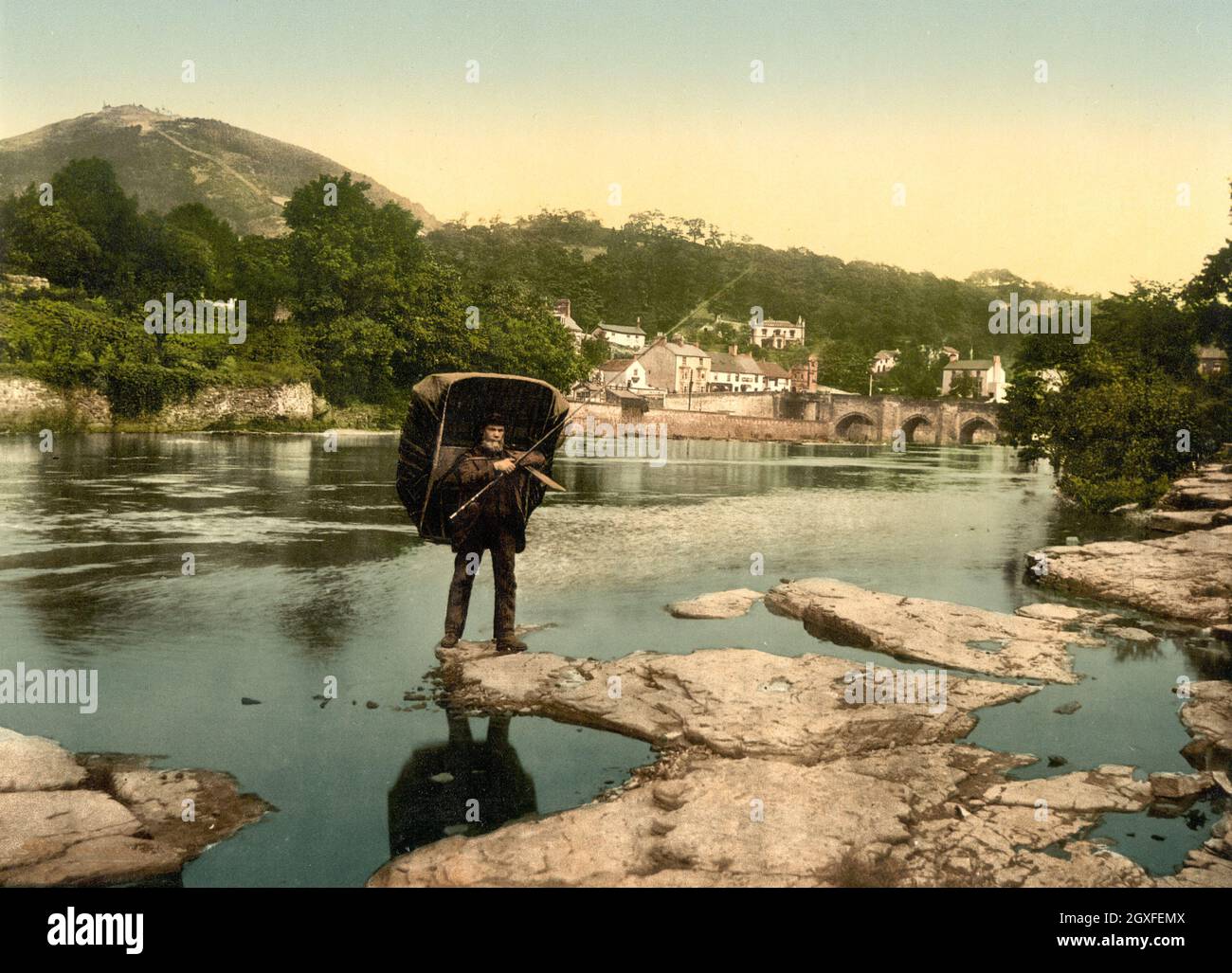 Image photochrome vintage de couleur main vers 1890 de la rivière Dee à Llangollen, au nord du pays de Galles, avec un pêcheur portant un bateau à coracle sur son dos. Le pont de Dee et la ville sont en arrière-plan avec le château de Dinas Bran sur la colline au-dessus de la ville. Banque D'Images