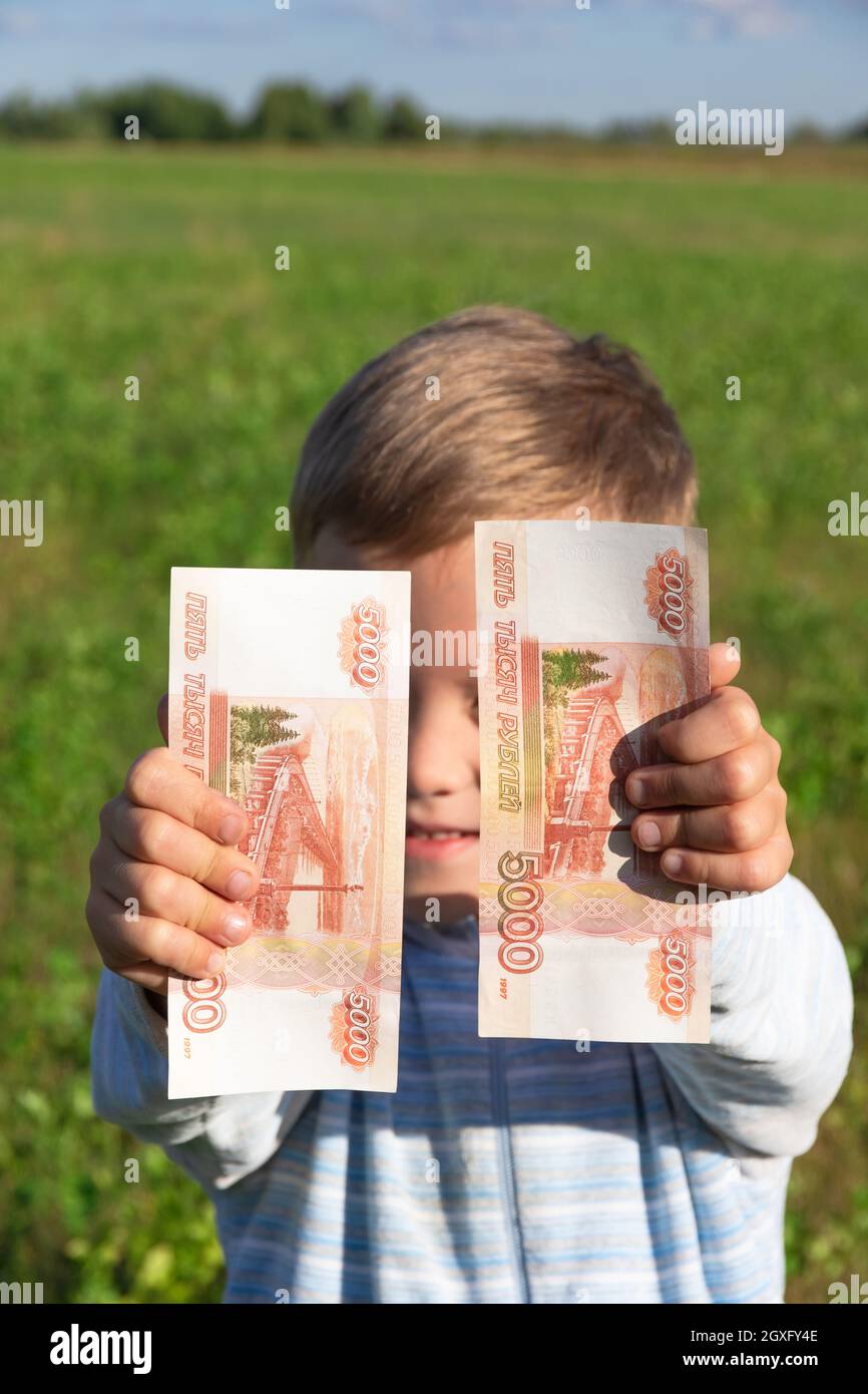 Un enfant satisfait, un garçon d'avant-garde, tient des roubles de papier dans ses mains à l'extérieur sur le fond d'un champ avec de l'herbe verte pendant un été ensoleillé Banque D'Images