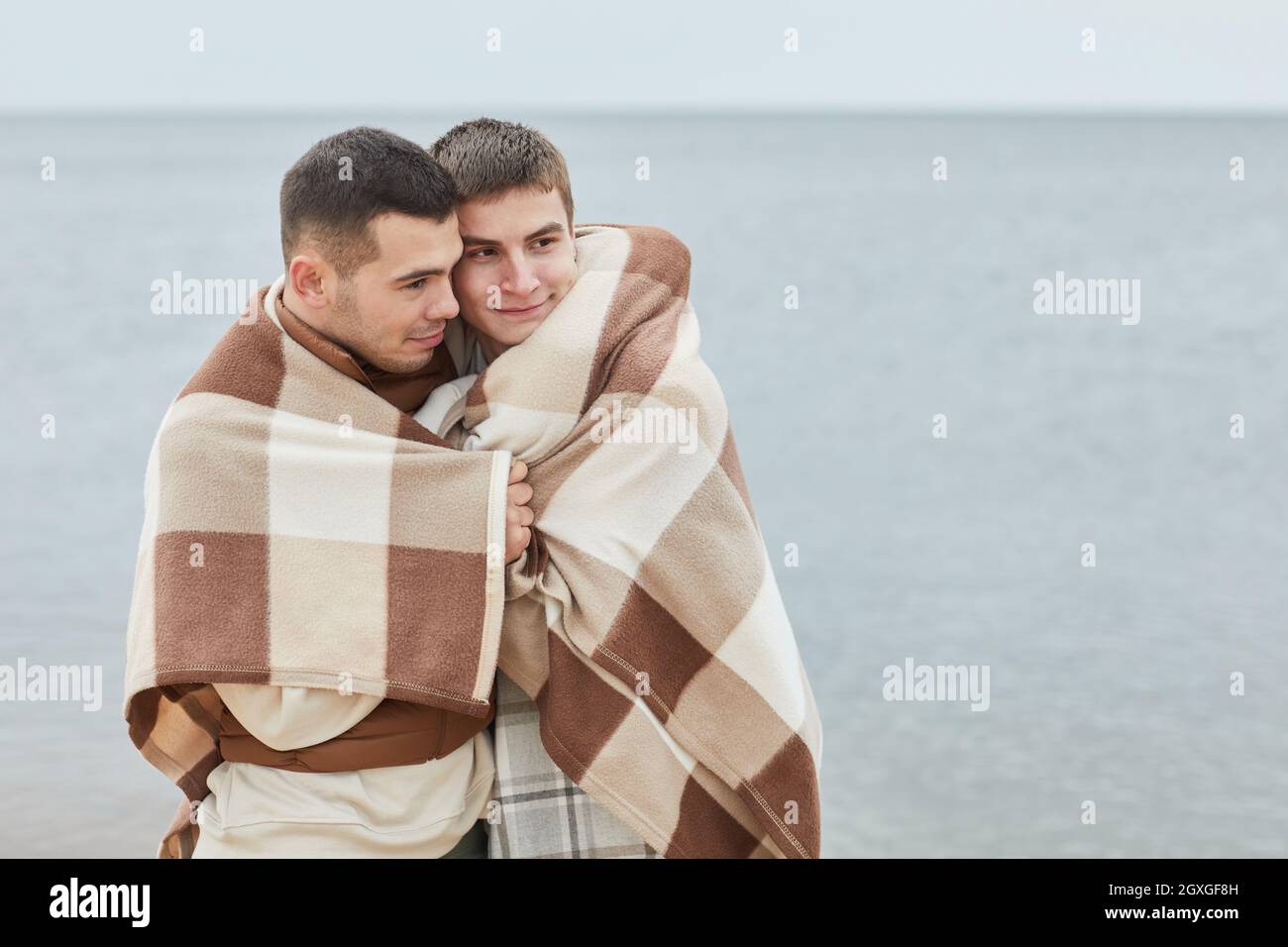 Portrait minimal d'un jeune couple gay embrassant sur la plage enveloppé dans une couverture avec de l'eau en arrière-plan, espace de copie Banque D'Images