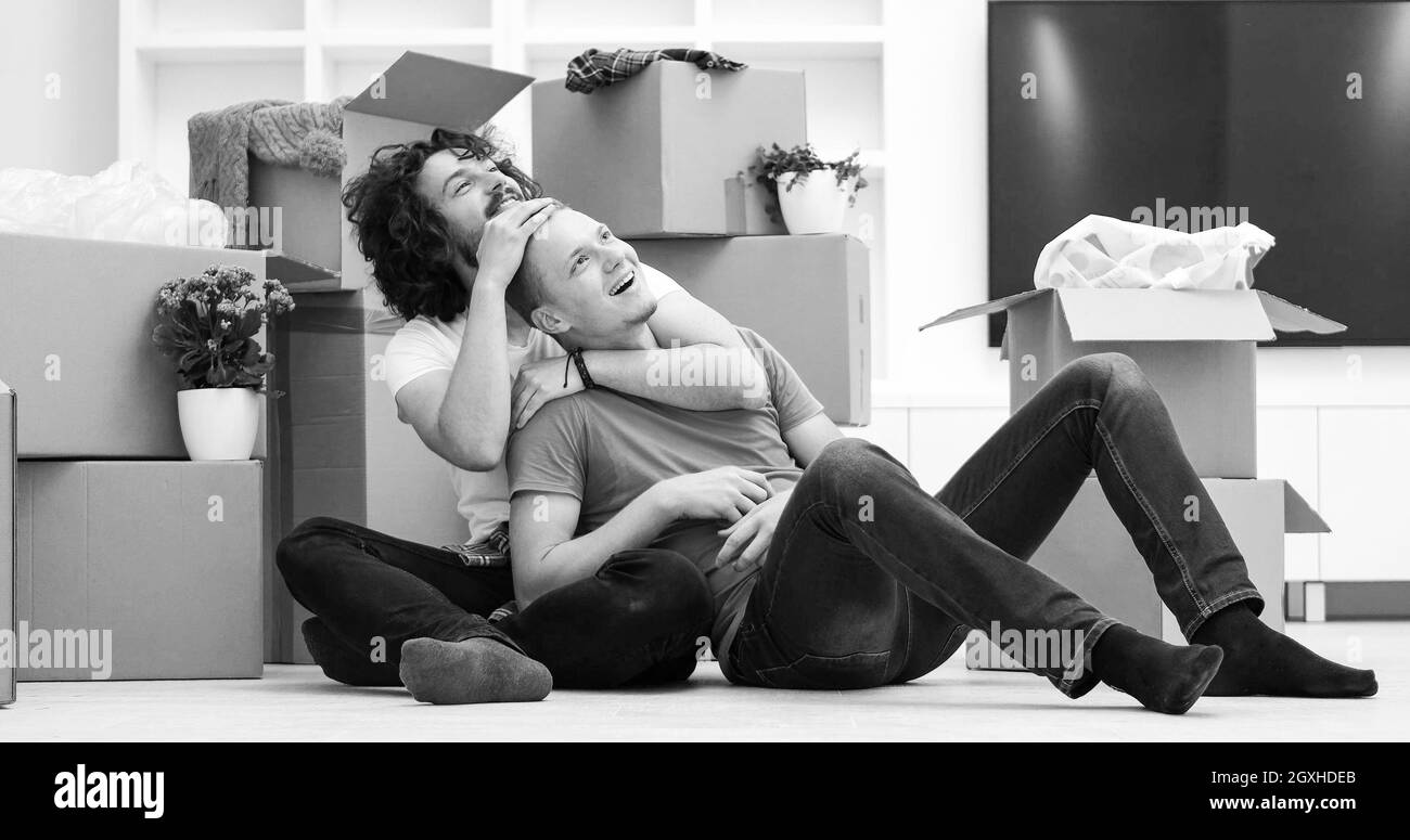 Se détendre dans maison neuve. Cheerful young gay couple assis sur le sol tandis que des boîtes en carton portant tout autour d'eux Banque D'Images