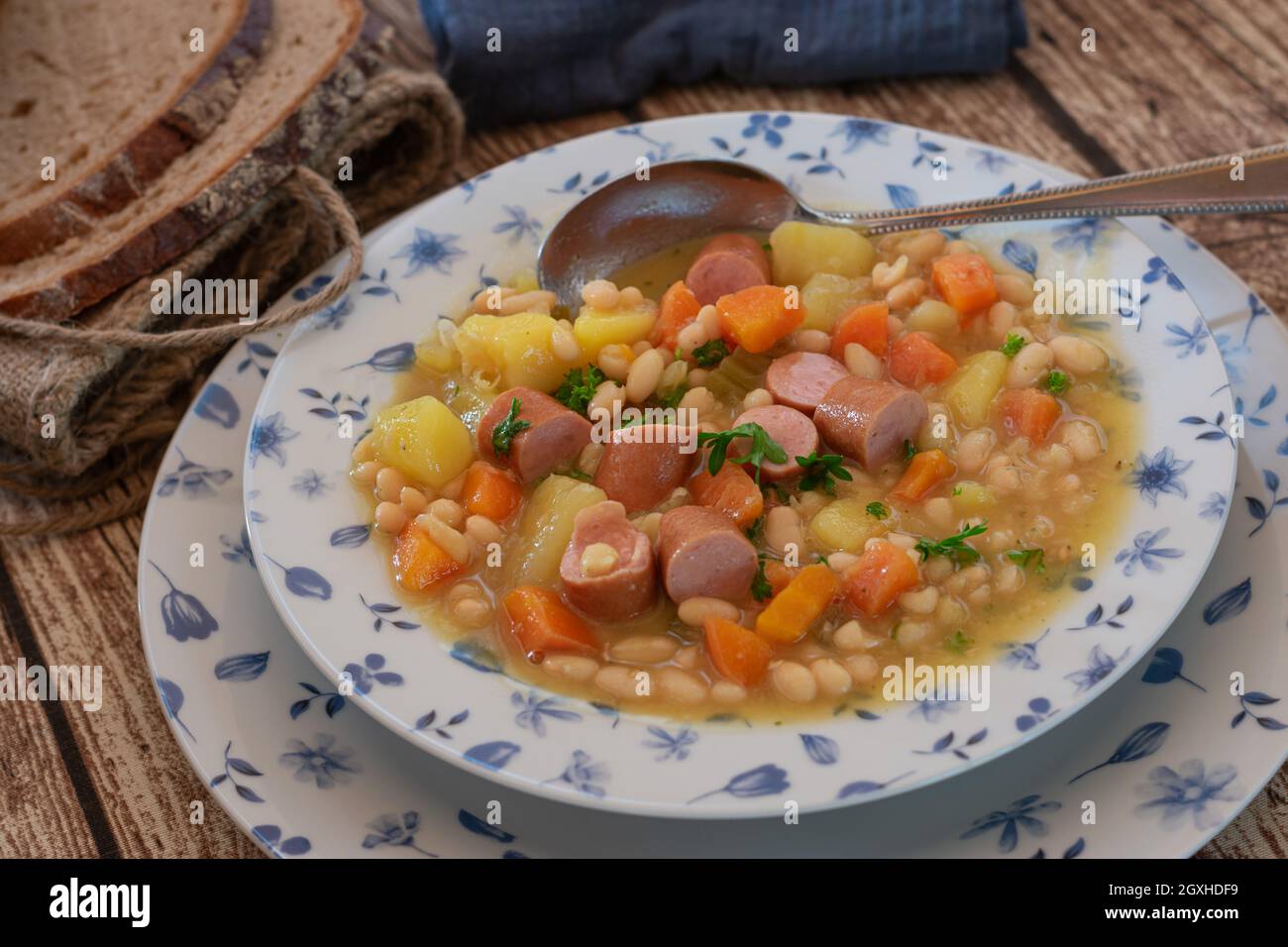 Ragoût de haricots blancs, légumes racines et saucisse de vienne.Servi sur une assiette rustique sur une table en bois avec une cuillère et du pain au levain. Banque D'Images